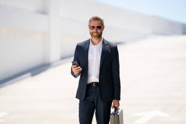İş portresi. Dışarıdaki telefondan iş adamı aradı. Başarılı lider hareket halinde. Takım elbiseli, çantalı bir adam telefonda konuşuyor. İşadamı işe gidiyor. Çantası olan bir iş adamı. İş başarısı