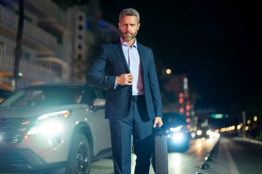 Gece vakti takım elbiseli bir iş adamı şehirde yürüyor. Takım elbiseli zengin bir adam. Lüks arabanın yanında bir iş adamı. Olgun bir iş adamı gece şehir ışıklarında yürür. İş stili. Zengin iş adamı