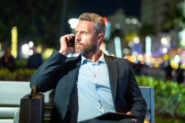 Takım elbiseli bir adam telefonda konuşuyor ve tablet kullanıyor. İş adamı dışarıyı arıyor. Tabletli ve telefonlu bir iş adamı. İş adamı aradı, bankta oturdu. Gece işi.