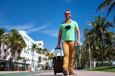 Bir gezgin Miami 'nin güneşli sokaklarında yürüyor. Bir erkek gezgin, tropik bir şehirde yaz tatilinin tadını çıkarır. Bavullu bir gezgin turist şehir manzarası. İnsan gezgin şehirde yürür