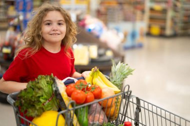 Süpermarkette el arabasıyla çocuk alışverişi. Çocuk markette taze sebze seçiyor. Süpermarkette yiyeceği olan bir çocuk. Süpermarkette ürün seçen bir çocuk. Süpermarkette taze yiyeceği olan çocuk