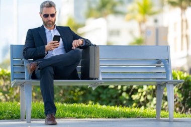 Kendine güvenen bir iş adamı bankta dizüstü bilgisayarı ve parktaki telefonuyla oturuyor. İspanyol iş adamı. Olgun iş adamı tatilin tadını çıkarıyor. İş adamı parktaki bankta mola veriyor. Yaz işleri.