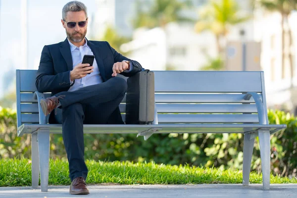 Kendine güvenen bir iş adamı bankta dizüstü bilgisayarı ve parktaki telefonuyla oturuyor. İspanyol iş adamı. Olgun iş adamı tatilin tadını çıkarıyor. İş adamı parktaki bankta mola veriyor. Yaz işleri.