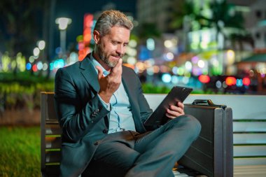 Takım elbiseli bir adam dışarıda tablet üzerinde çalışıyor. Tabletli bir iş adamı. Gece sokağında tabletli bir iş adamı. Tablet kullanan bir işadamı, bankta oturur. Gece işi.