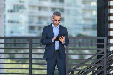 Açık havada tabletle başarılı bir iş adamı. Kendine güvenen bir iş adamı dışarıda tablet kullanıyor. Olgun iş adamı girişimci finans verilerini kontrol eder. Zengin bankacı tabletteki sözleşmeyi gözden geçiriyor