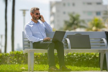 Açık havada dizüstü bilgisayar üzerinde çalışan başarılı bir iş adamı. Kendine güvenen bir iş adamı elinde dizüstü bilgisayarla bankta oturuyor. İspanyol işadamı uzaktan çalışıyor. Yazın internette çalışan zengin iş adamı