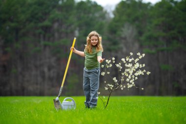 Çocuk dışarıda ağaç dikiyor. Bitkilerin bahar etkinliği. Çocuk kürek ve su bidonuyla çalışıyor. Çocuk meyve ağacı dikmiş. Bahçıvan çocuklar. Çocuk ağaç dikmek ve onunla ilgilenmek. Bahar otlağında küçük çiftçi