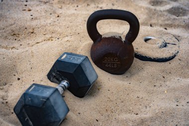Kettlebell ve ağırlık arka planı. Ağır ağırlıklarla vücut geliştirme. Dambıllarla park egzersizi. Ağırlıklarla spor sahnesi. Kettlebell, halterler ve halterler. Kumsalda ağırlık çalışması.