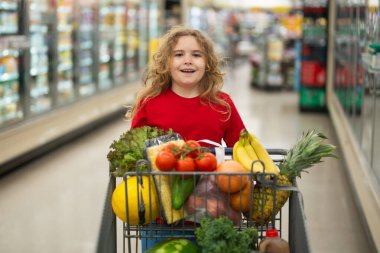 Süpermarkette el arabasıyla alışveriş yapan çocuk. Çocuk markette taze sebze seçiyor. Yiyecek satan bir çocuk. Markette ürün seçen bir çocuk. Bakkalda taze yiyeceği olan bir çocuk.