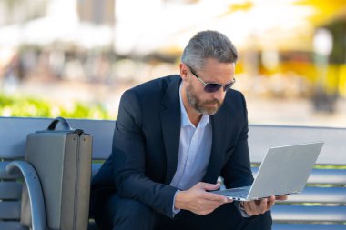 Kendine güvenen işadamı bankta dizüstü bilgisayarı ve parktaki telefonuyla oturuyor. Başarılı bir iş adamı. Olgun iş adamı tatilin tadını çıkarıyor. İş adamı parktaki bankta mola veriyor. Yaz işleri.