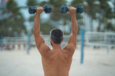 Spor adamının dambılları açık havada. Spor aktivitelerine katılan atletik bir adam. Formda adam geri döndü. Spor egzersizleri. Spor seansındaki üstsüz adam, geri döndü. Dambıllı açık hava eğitimi