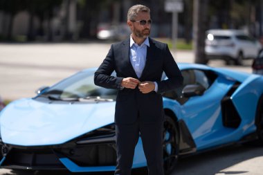 Olgun iş adamı. Zengin iş adamı başarının tadını çıkarıyor. Lüks bir iş. Pahalı arabanın önündeki zengin adam. Lüks iş seyahati. Klasik takım elbiseli, kendinden emin zengin bir iş adamı.