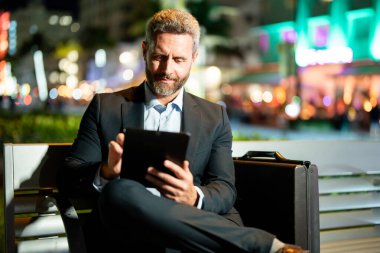 İnternet işi. Dışarıda tablet üzerinde çalışan bir iş adamı. Tabletli bir iş adamı. Gece sokağında tabletli bir iş adamı. Tablet kullanan bir işadamı, bankta oturur. Gece işi. Uzak çalışma