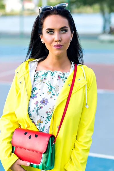 Attractive woman wears yellow clothes posing at basketball court on blur background. Outdoor photo of interested brunette girl with trendy bag waiting for someone before training.