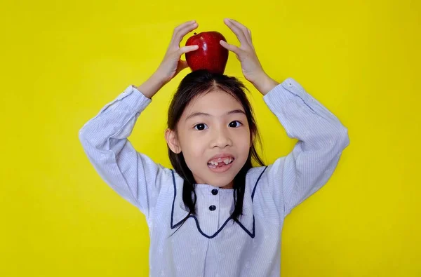 Kafasının üstünde kırmızı bir elmayı dengelemeye çalışan tatlı bir Asyalı kız, onu bırakmadan önce elleriyle beşiyormuş. Parlak sarı arkaplan.