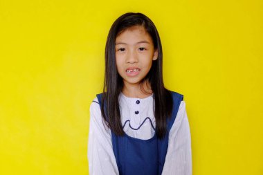 Beyaz ve mavi okul üniformalı, uzun saçlı, gülümseyen parlak, mutlu ve çekici bir Asyalı kızın yarı vücut fotoğrafı. Parlak sarı arkaplan.