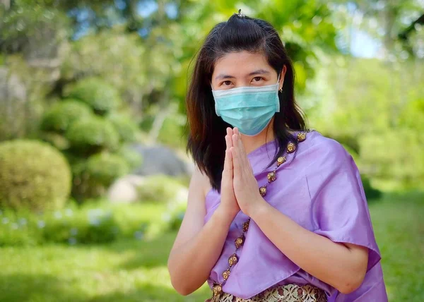 Geleneksel Tayland kostümü giyen ve cerrahi maske takan Tayland 'lı bir kadın ellerini öne eğip gülümseyerek & quot; Wai & quot;' u selamlıyor. El sıkışmak için bir karşılama.
