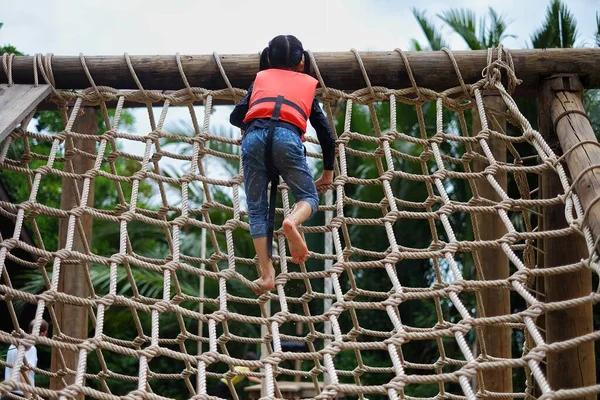 Turuncu can yeleği giyen genç bir Asyalı kız yaz kampında engelli parkurda tırmanıyor..