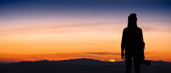 Back view of young hipster photographer holding the camera with sunrise on mountain natural background.