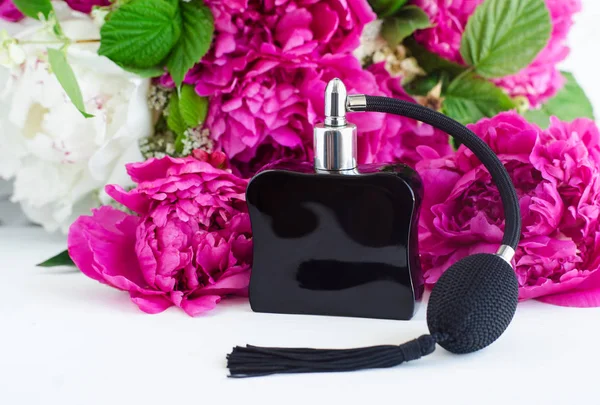 Black vintage perfume bottle with atomizer on the white table. Peony flowers blossom background. Copy space.