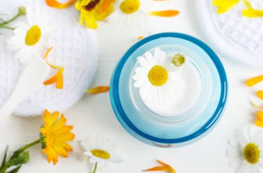 Mavi ışıklı cam kavanoz beyaz yüz kremi/maske ile. Beyaz arka plan üzerinde papatya ve calendula çiçekler. Üstten görünüm, kopya alanı.  