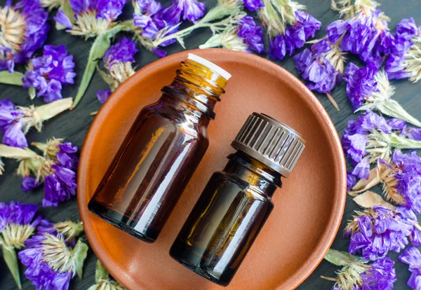 Two small bottles with essential oils on the small ceramic plate. Dark wooden background with dry purple flowers. Aromatherapy, spa and herbal medicine concept. Copy space, top view.