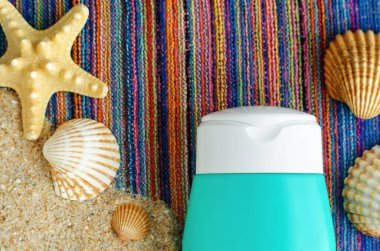 Kum ve kabuklu renkli plaj havlusunun üzerinde güneş kremi olan küçük bir şişe. Yaz cilt bakımı ve güneş koruma konsepti. Kapat, üst görünüm, boşluğu kopyala 