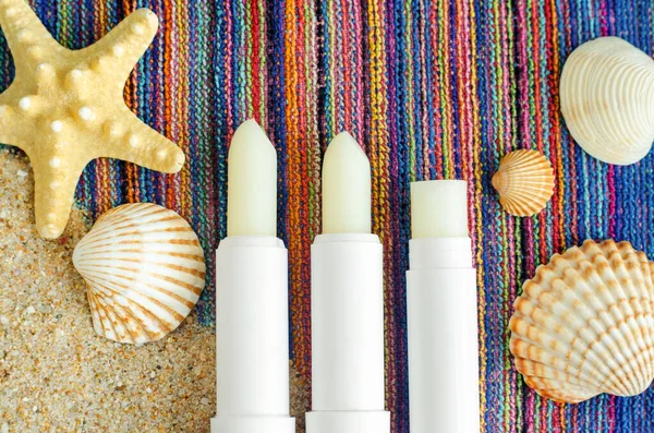 Three lip balms, sunscreen sticks on the colorful beach towel with sand and seashells. Summer lip treatment and UV protection concept. Close up, flat lay, copy space