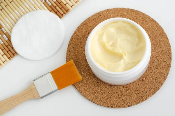 Yellow facial mask (banana face cream, hair mask, body shea butter) in the small white jar and make-up brush. Natural skin and hair concept. Top view, copy space.