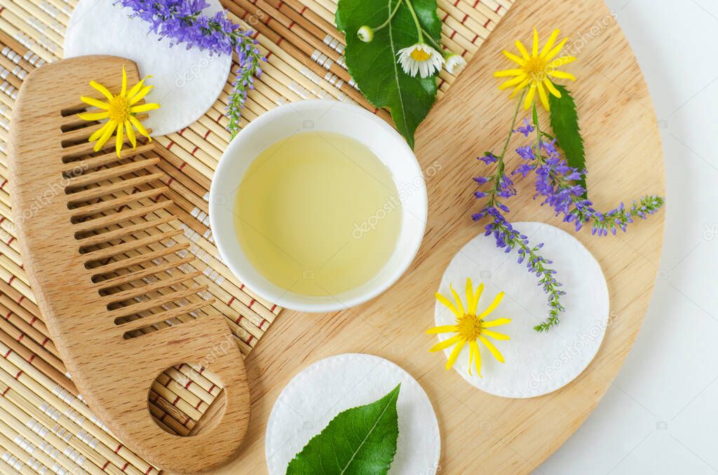 Small white bowl with cosmetic (massage, cleansing) aroma oil, wooden hair brush, cotton pads and wild flowers. Natural skin care, homemade spa and beauty treatment recipe. Top view, copy space. 