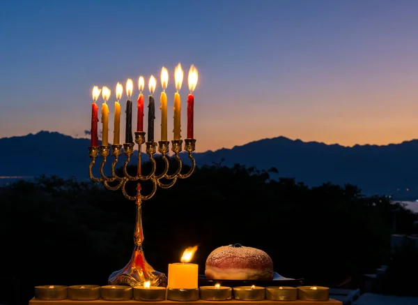 Hanuka için şenlikli bir kompozisyon, menorah yanan mumlar fener ve karanlık gökyüzünde tatlı çörekler ve dağ arka planıyla.