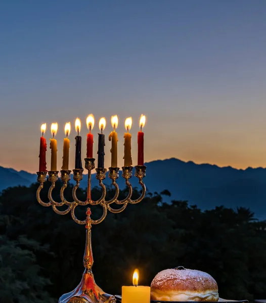 Menorah mumları, fener ve akşam gökyüzünde tatlı çöreklerle ve arka planda dağlarla Hanukkah için şenlikli bir kompozisyon.