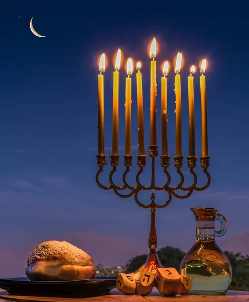 Sweet donuts, festive menorah with burning candles, jar with olive oil and wooden dreidels of teetotum. Each side of that bears Hebrew letter that together in English means - a great miracle happened here, background of clouds above mountains