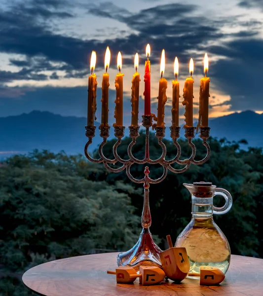 Sweet donuts, festive menorah with burning candles, jar with olive oil and wooden dreidels of teetotum. Each side of that bears Hebrew letter that together in English means - a great miracle happened here, background of clouds above Sinai mountains