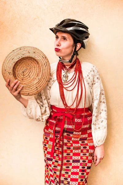 beautiful cheerful girl in traditional Ukrainian clothes (embroidery), an ancient chain, necklaces and a modern bicycle helmet and a straw hat in their hands (steering wheel, wheel, change of priorities, turning in life - concept)