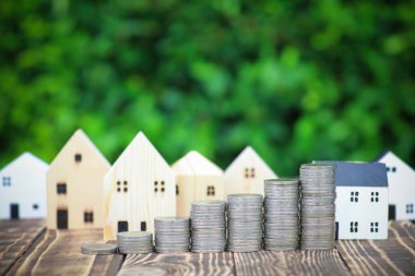 Arkasında ağaç yeşili bulanık arka planı olan tahta masaya ve model eve bozuk para yığmak. Gelecekteki konut alım-satımı ve iş yeri yatırımları için para biriktirme kavramı.