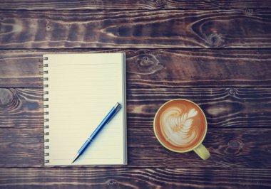 Üst manzara, sarı bir fincanda düz kahve, boş bir defter ve eski ahşap masa zemininde fotokopi alanı olan mavi bir kalem..