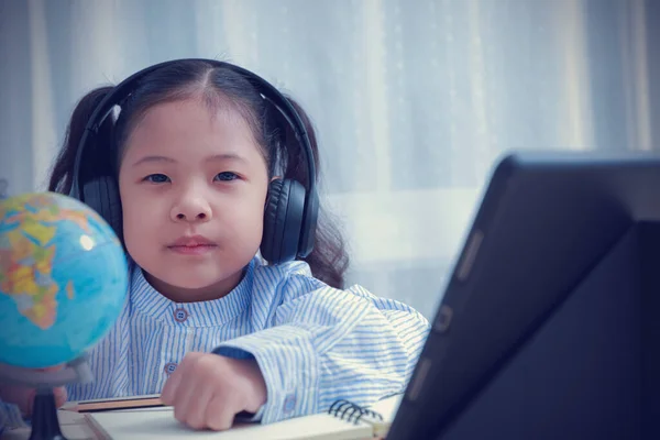 Asian kid girl wearing headphones looking at the camera learning education distance from home by tablet. Current kids around the world learning distance from home during the coronavirus outbreak using a long time.