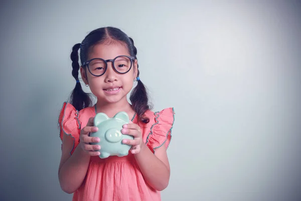 Asyalı küçük kız, elinde bir kumbarayla gelecek için para biriktirirken gülümsüyor. Tasarruf, bankacılık, muhasebe kavramı.