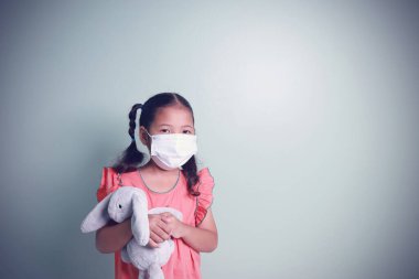 Korunma virüsü ve toz için beyaz maske takan Asyalı şirin kız portresi yeşil arka planda fotokopi alanı olan tavşan bebeğine sarılıyor..