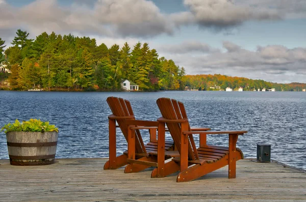 İki Muskoka sandalyesi arka planda ağaçlar ve kulübeler olan sakin bir göle bakan bir rıhtımda oturuyor.