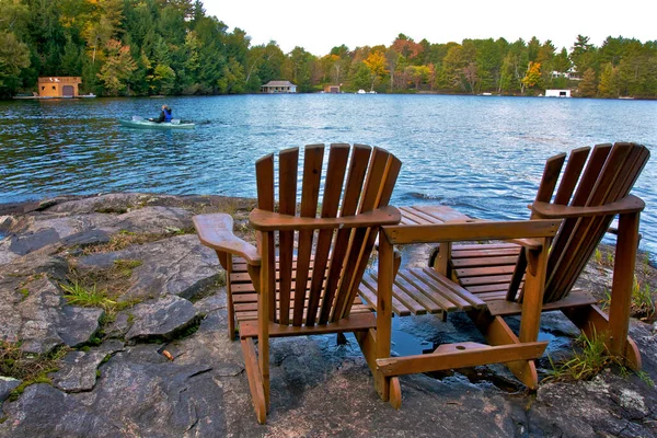 Kayalık bir kıyıda oturan iki Muskoka sandalyesi arka planda kulübeleri olan sakin bir göle bakıyor.