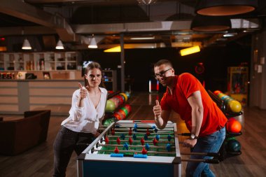 Güzel bir kız ve gözlüklü bir adam oyun odasında futbol oynuyorlar ve başparmak kaldırıyorlar. Aşk ve eğlence.