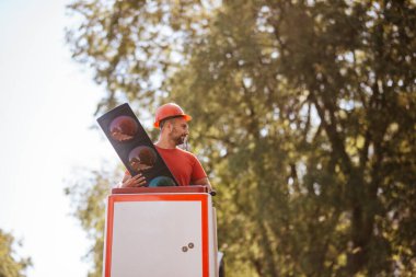 Kırmızı tişörtlü beyaz bir işçi elinde trafik ışığı olan bir vinçteki sepetin içinde duruyor. Trafik ışıkları ayarlanıyor