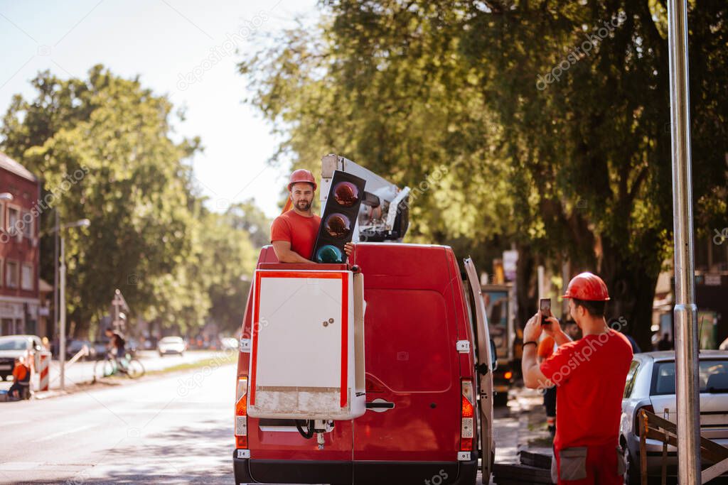 A caucasian worker in a red T-shirt stands in a basket in a crane with a traffic light in his hands. Setting up traffic lights in progress