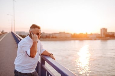 Beyaz gömlekli, gözlüklü orta yaşlı bir işadamı, telefonda konuşuyor, köprüye yaslanıyor.