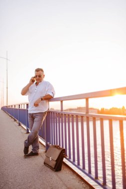 Beyaz gömlekli, orta yaşlı, beyaz gözlüklü beyaz bir işadamı telefonda köprü parmaklıklarına yaslanmış konuşuyor.