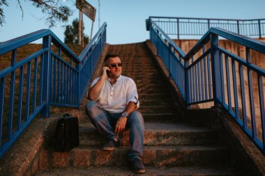 Gözlüklü ve beyaz gömlekli orta yaşlı bir işadamı merdivenlerde oturup telefonda konuşur.