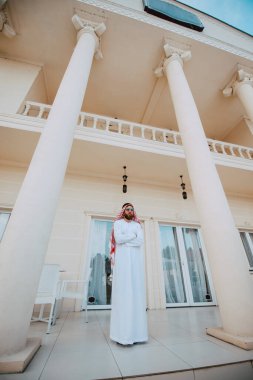 Geleneksel kıyafetli yakışıklı orta doğulu Müslüman adam beyaz, güzel, modern evin önünde duruyor.