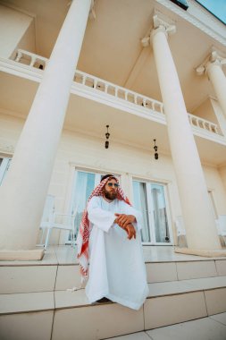 Geleneksel kıyafetli yakışıklı orta doğulu Müslüman adam beyaz, güzel, modern evin önünde duruyor.